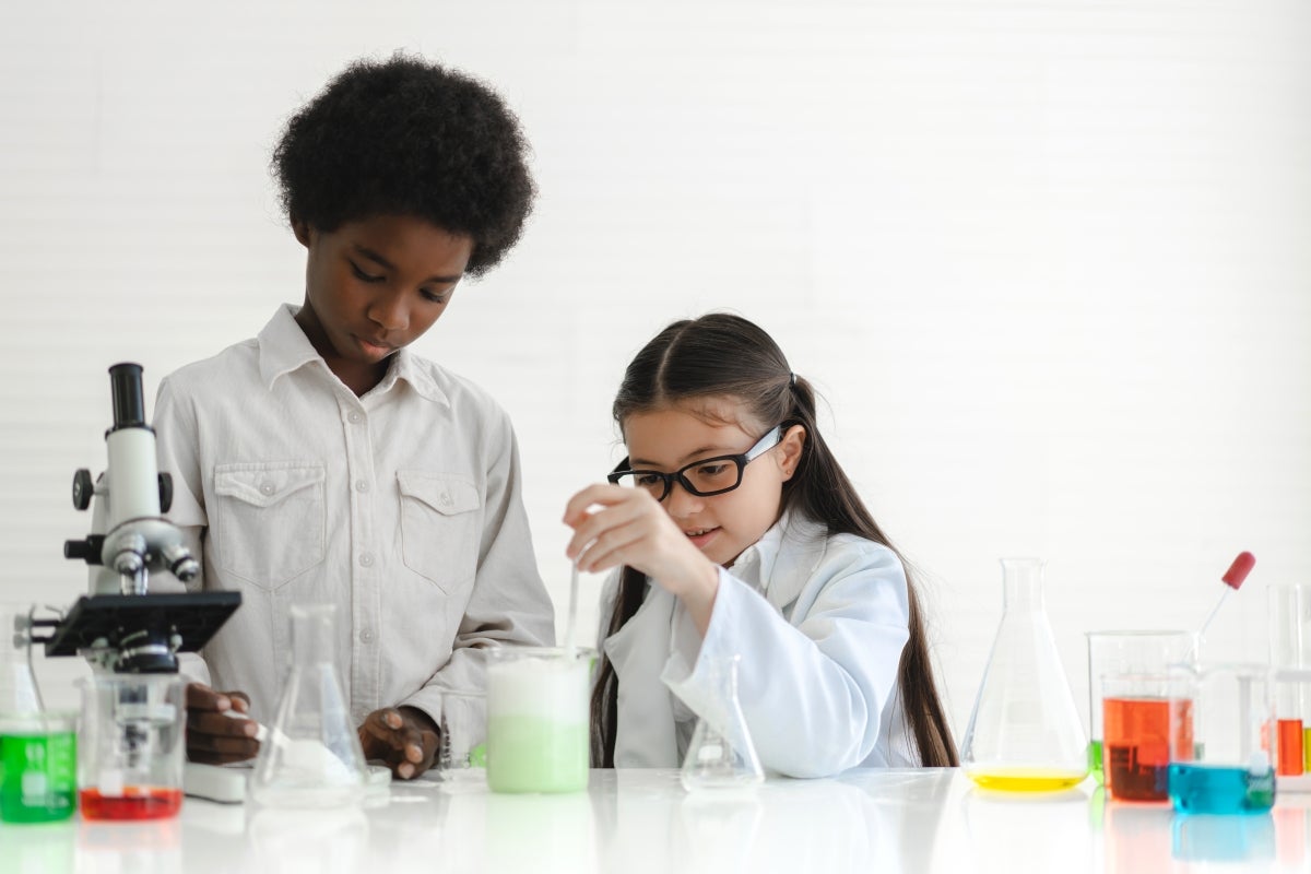 In a laboratory two youth scientists wear white lab coats and are researching chemicals with glass beakers filled with colorful liquids.